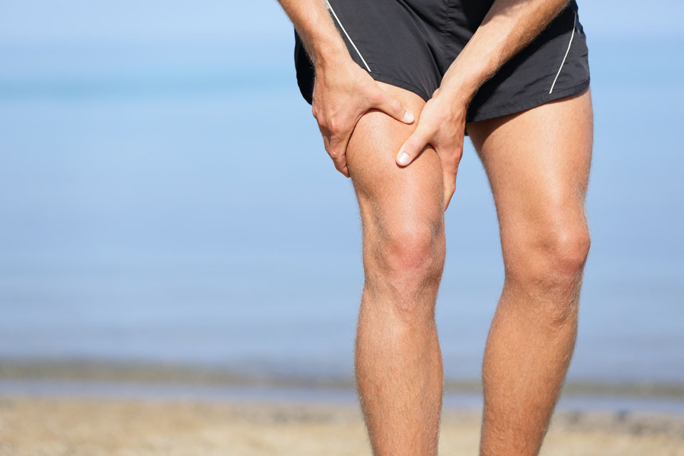 Muscle injury. Man with sprain thigh muscles. Athlete in sports shorts clutching his thigh muscles after pulling or straining them while jogging on the beach.