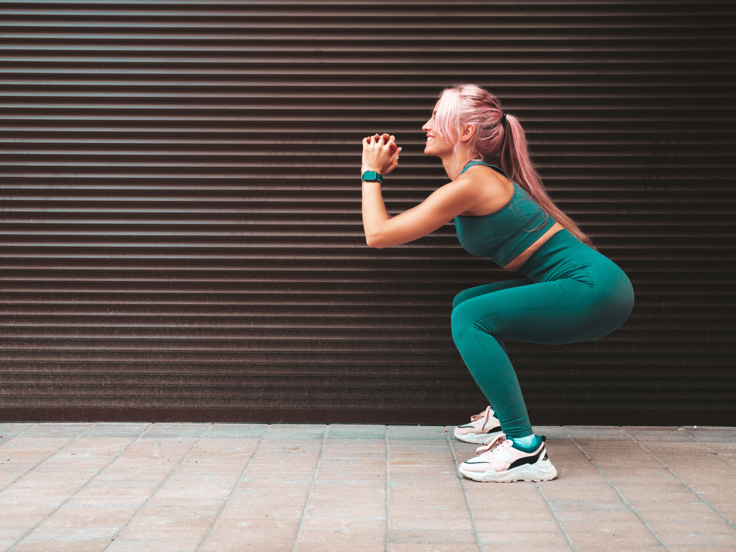 Sexy fitness woman in green sports clothing with pink hair.She doing squats. Young beautiful model with perfect body.Female in the street near roller shutter wall.Cheerful and happy. Outdoors
