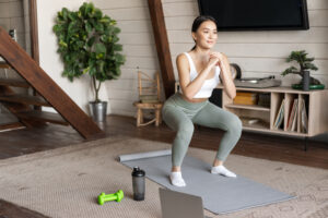 Young asian fitness girl doing squats workout at home, follow online sport gym instructor, standing on floor mat and smiling, exercising in living room Young asian fitness girl doing squats workout at home, follow online sport gym instructor, standing on floor mat and smiling, exercising in living room.